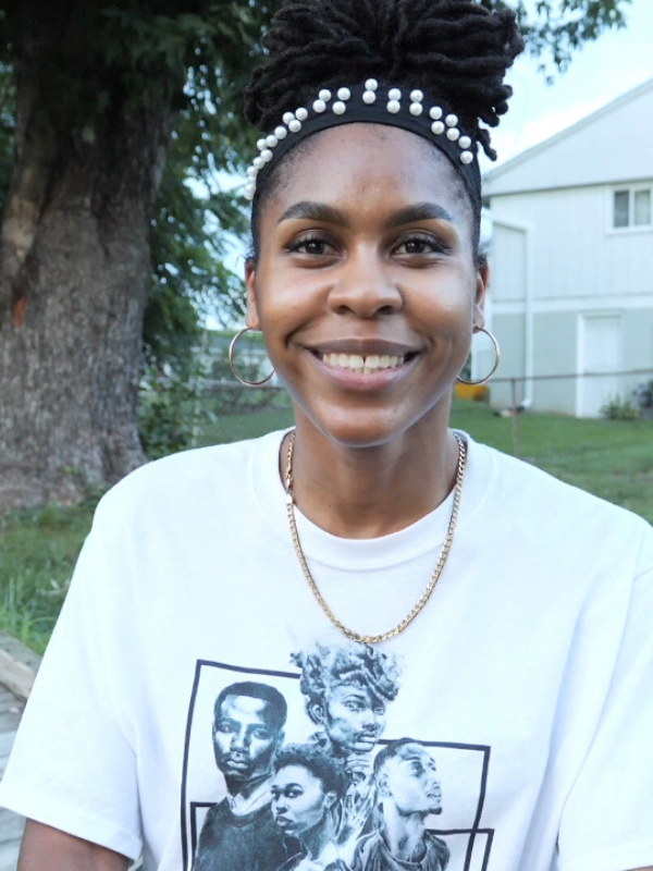 Tyree Brown, a Black woman is smiling at the camera. She is wearing a black headband with pearls with her hair tied up, hoop earrings, and a white tshirt.