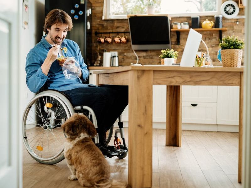 Man in wheelchair eats a meal while interacting with his dog