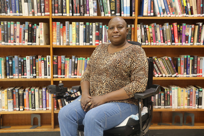 Francene, a beautiful bald, Black woman, sits in an electric wheelchair and gazes gently forward. She’s wearing a leopard print long sleeve shirt and blue jeans, and sits in front of a full bookshelf.