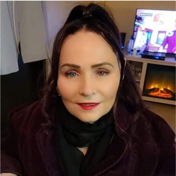 Shannon, a white woman with dyed purple/black hair smiles directly to camera. She wears a black scarf and top and pink lipstick.