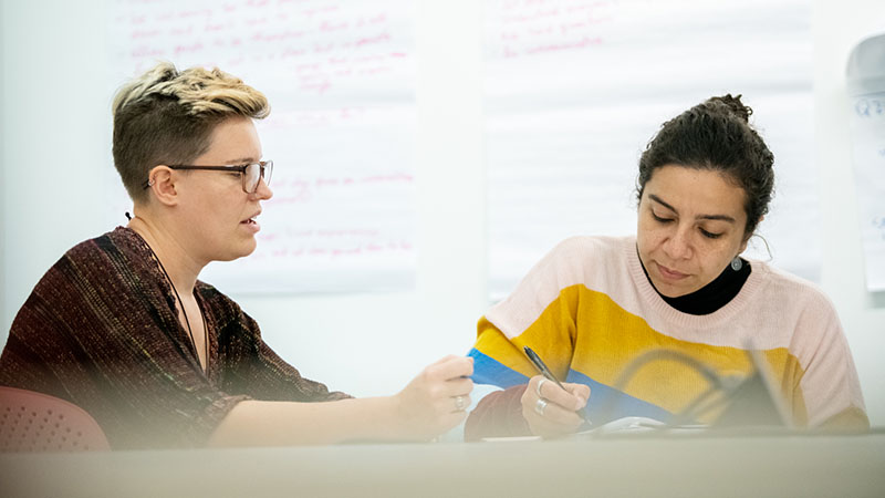 Two students working together at a table in front of a whiteboard