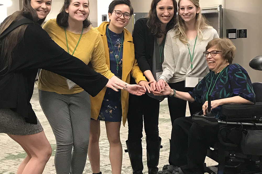 5 undergraduate fellows are pictured standing in a semicircle around Judy Heumann