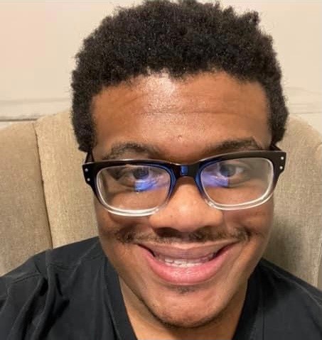 A smiling African-American man, sitting in a chair. He has an Afro, glasses, mustache, and a black t-shirt.