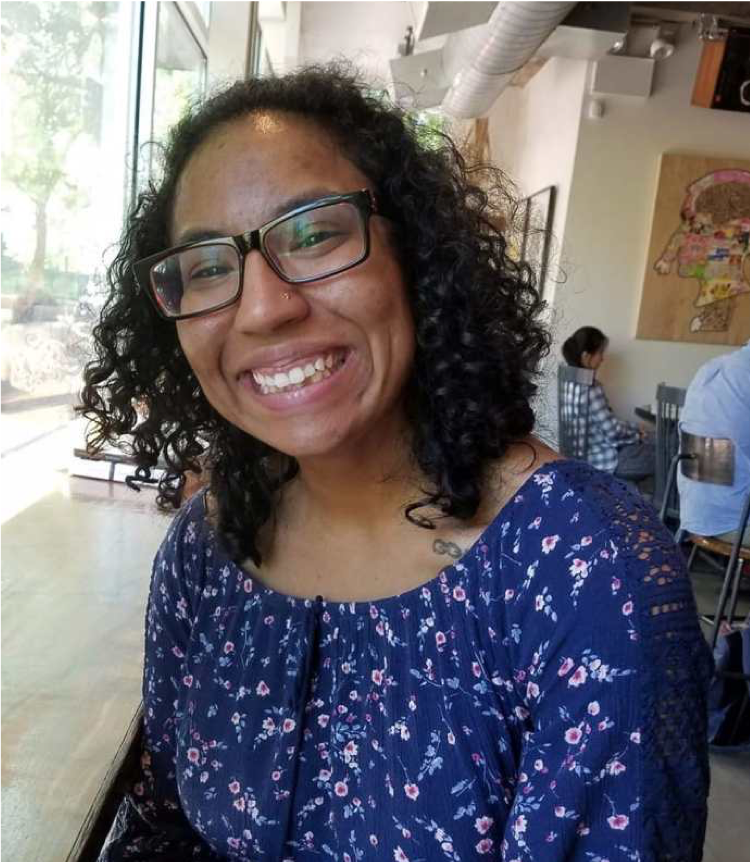 Valerie is a Black and Latina person with chin length, curly black hair. They are wearing black, square glasses and a blue, flowered top. They are sitting  in a coffee shop smiling towards the camera 