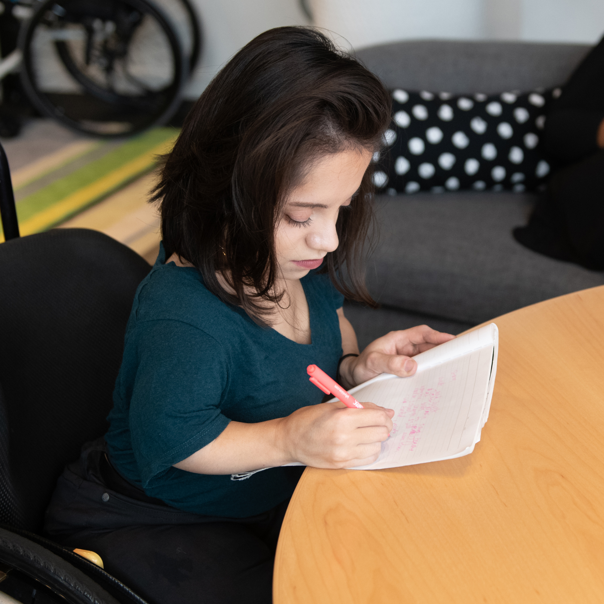 A South Asian person in her wheelchair takes notes by hand during a meeting.