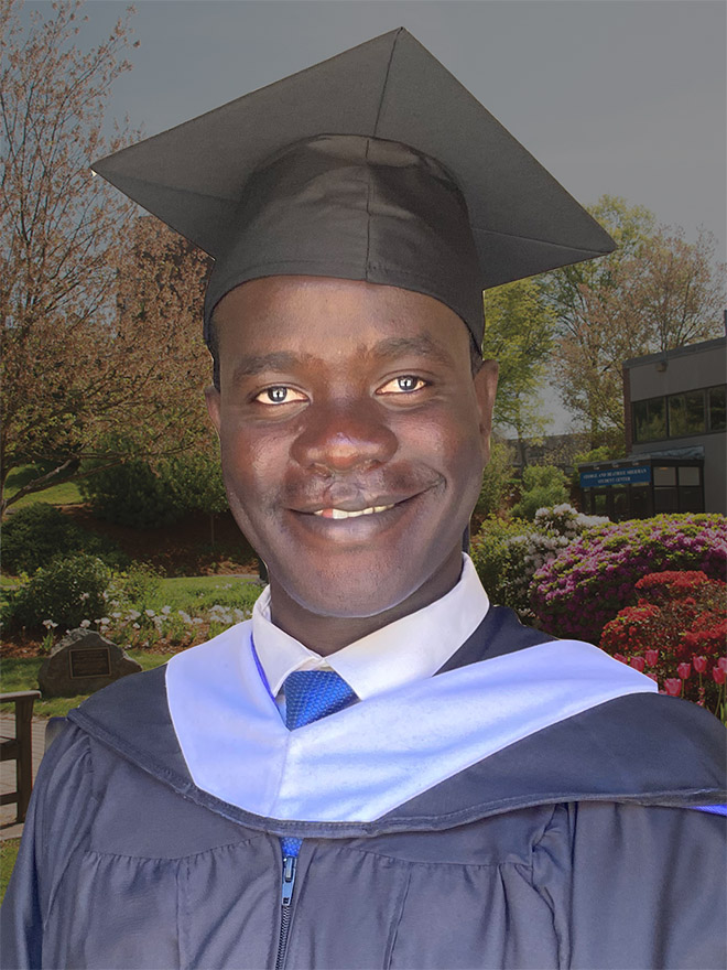 Francis Ojok, MA COEX'21, in a commencement cap and gown in front of flowers, a bench, tree and building at Brandeis