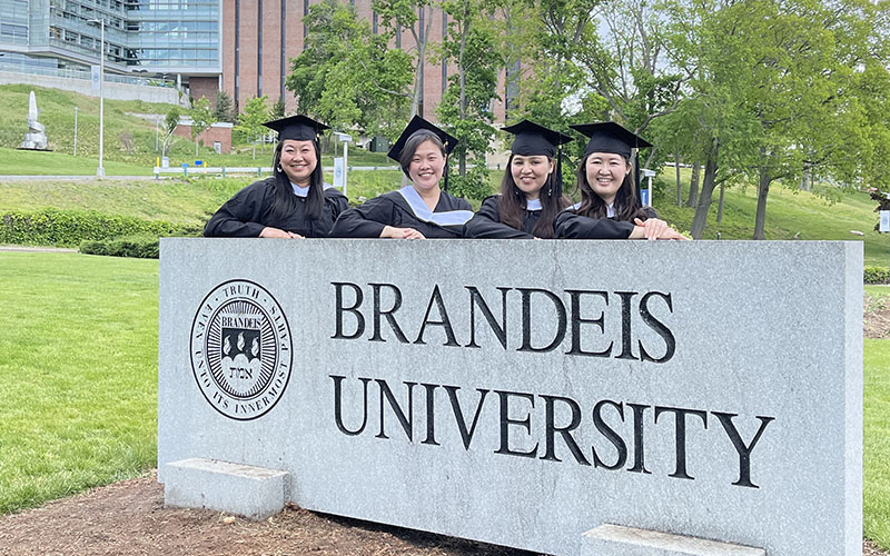 Four Heller students in graduation regalia behind the Brandeis University stone sign