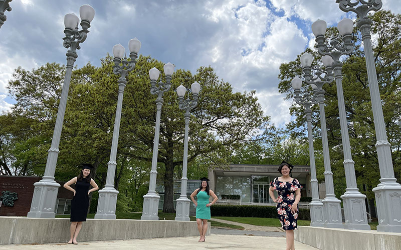 Three Heller students in dresses and graduation caps pose with street lamps outside Rose Art Museum
