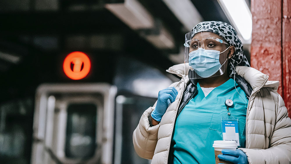 A Black nurse wearing a face mask, face shield, scrubs and coat waiting for a train