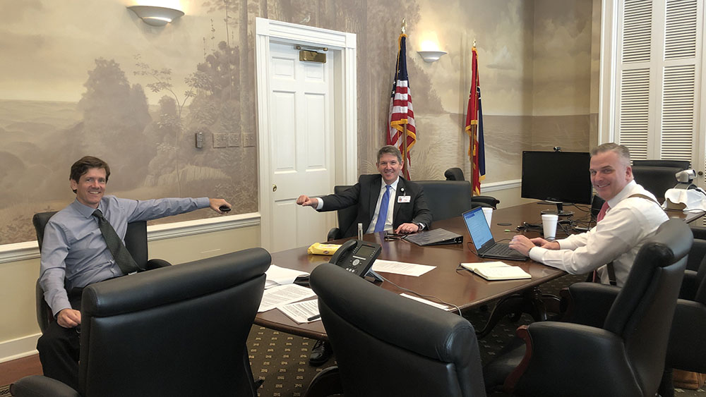 Mississippi State Health Officer Dr. Thomas Dobbs, Dr. Clay Hays and David Maron, chief legal counsel to Gov. Tate Reeves, around a wooden table with flags in the background