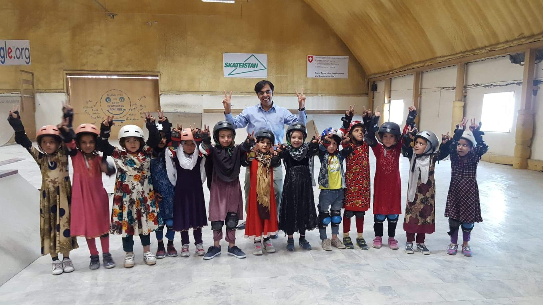 Soroush Kazemi with young girls in helmets and elbow pads, putting their arms up with peace signs, at Skateistan