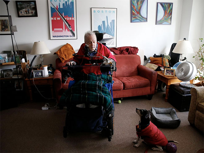 A man in a wheelchair in his home, covered in blankets, looking at a dog