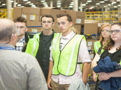 Workers in bright green vests face a man in a collared shirt standing in a warehouse