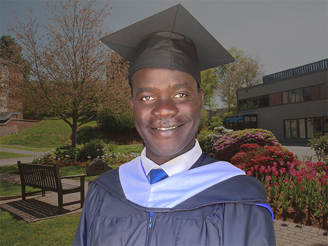 Francis Ojok, MA COEX'21, in a commencement cap and gown in front of flowers, a bench, tree and building at Brandeis