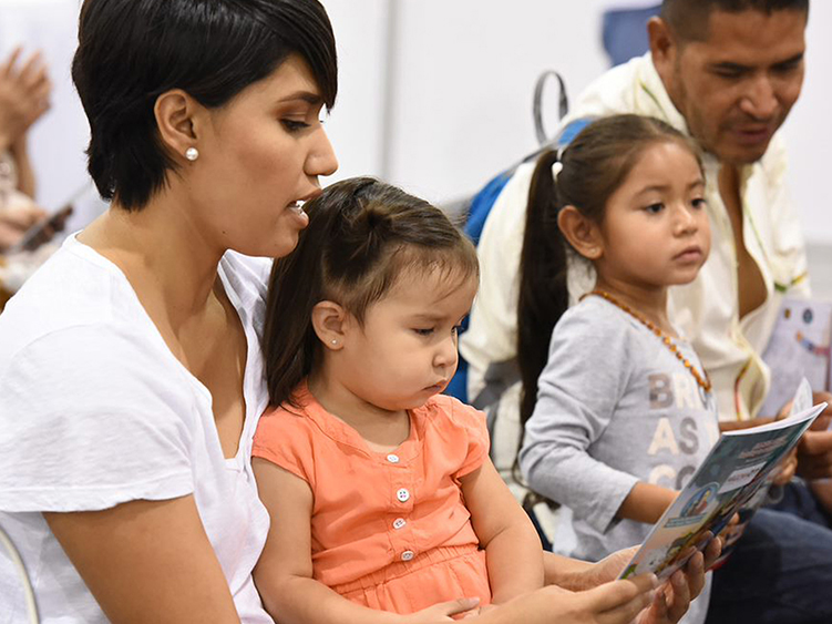 A woman reads to a child with other children and adults in the background