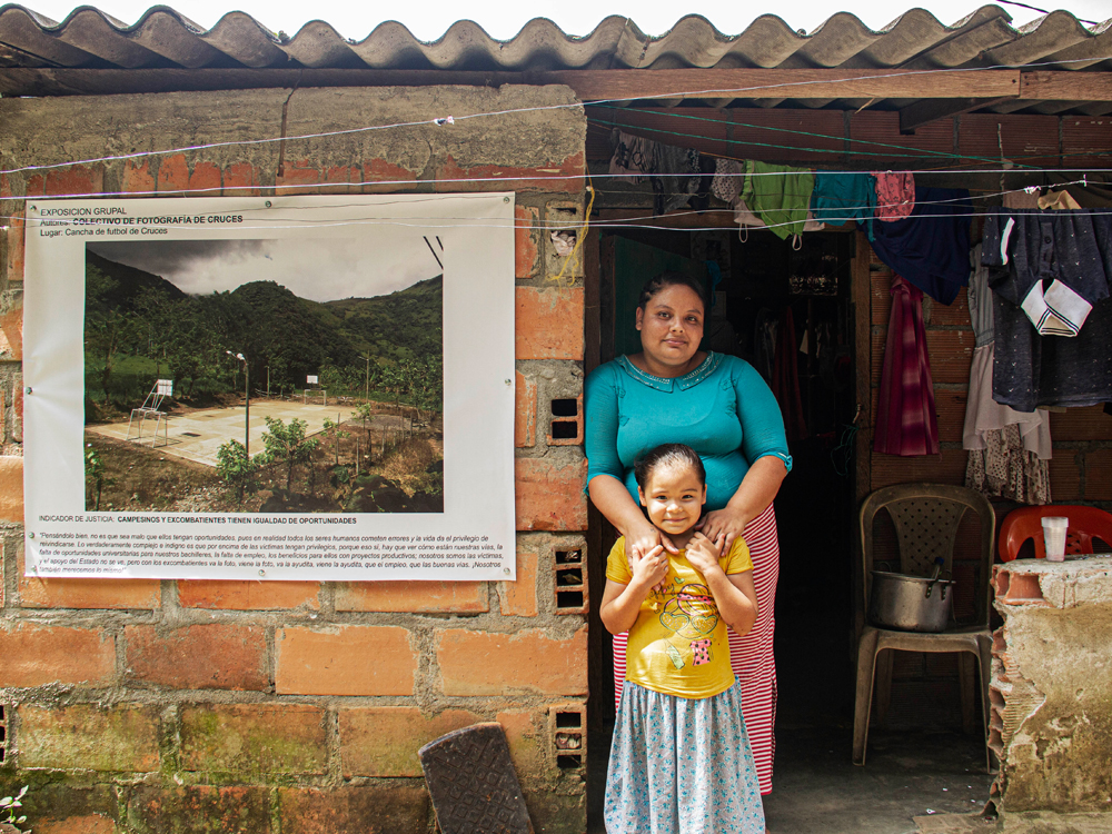 A woman and a child stand next to a poster depicting a photo of a vacant soccer field, in Cruces, Colombia.