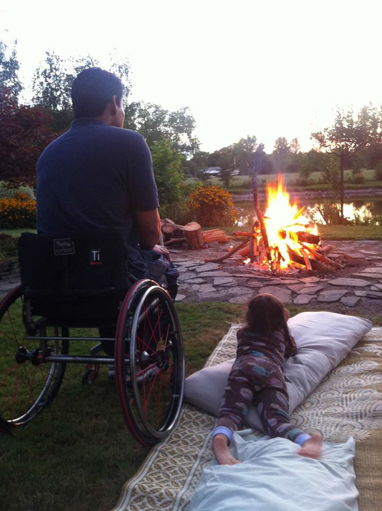 A father using a wheelchair and his daughter laying on a blanket sit near a campfire.