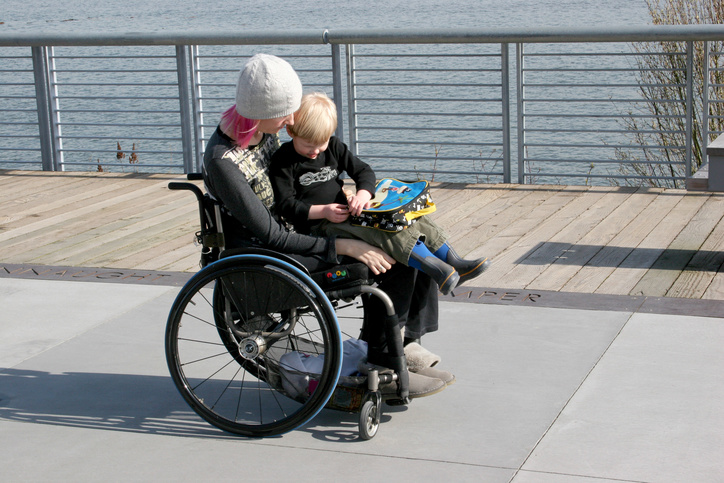 A mother using a wheelchair alongside the river with her young child holding his backpack on her lap