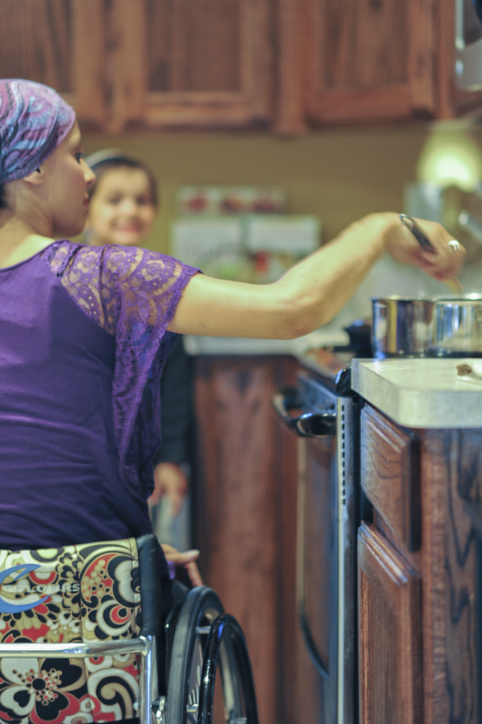 A mother using a wheelchair stirs a pot on the stove while her child stands nearby