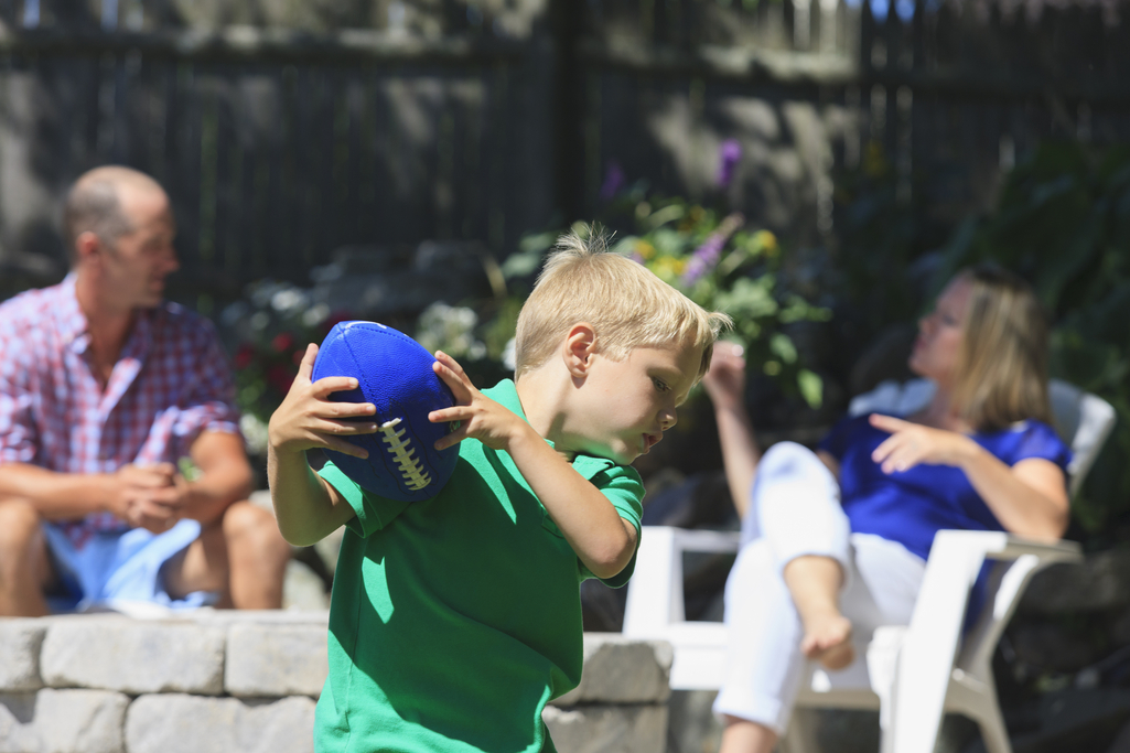 A young boy plays catch with a football while his parents are sitting nearby.