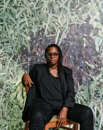 Mickalene Thomas in an all black suit, sitting in front a wall of green leaves.
