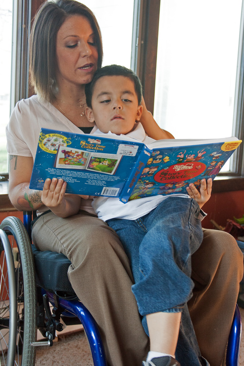 A mother using a wheelchair reads a book to her son while he sits on her lap