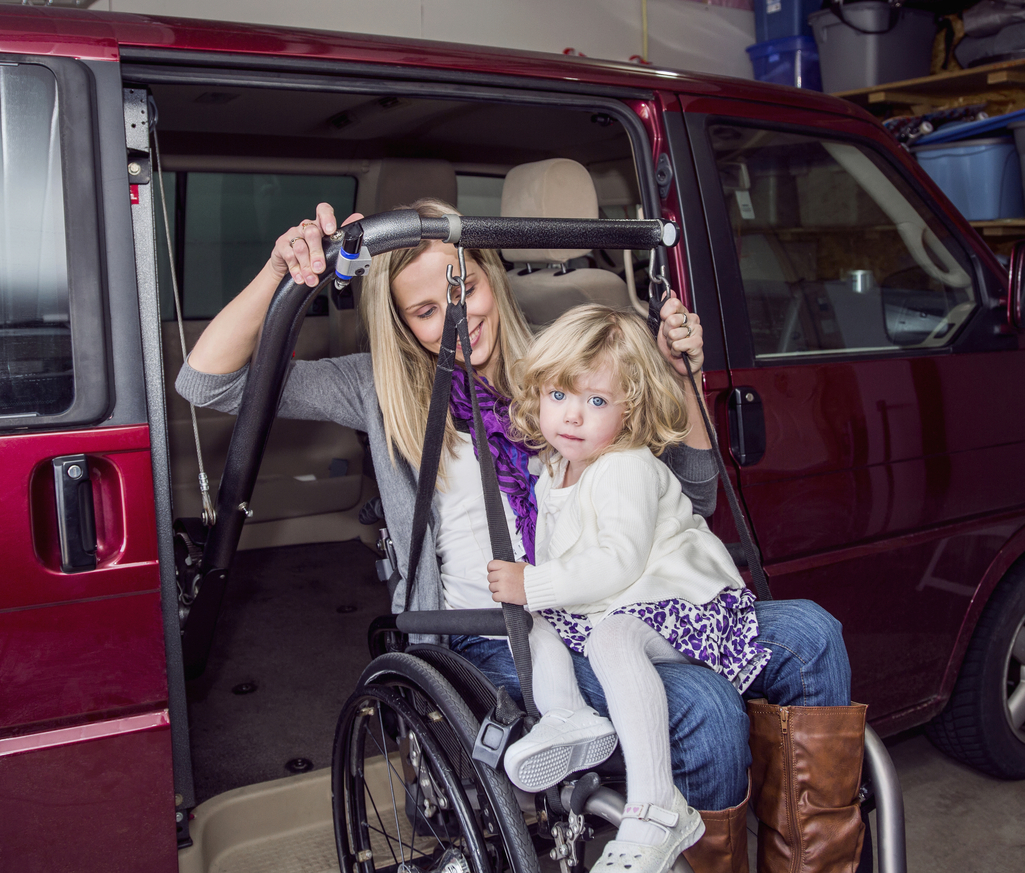 A young girl sits in her mother's lap. Her mother is in her wheelchair and they are entering her van.