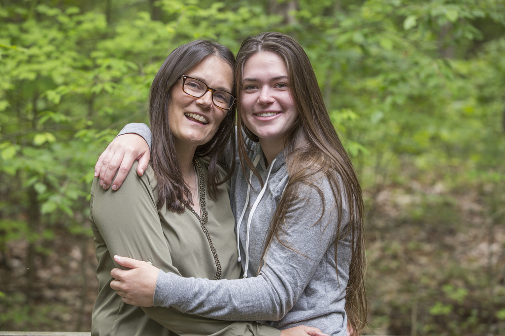 Una madre y su hija adolescente o adulta joven están de pie, abrazadas, y detrás de ellas hay un fondo de árboles