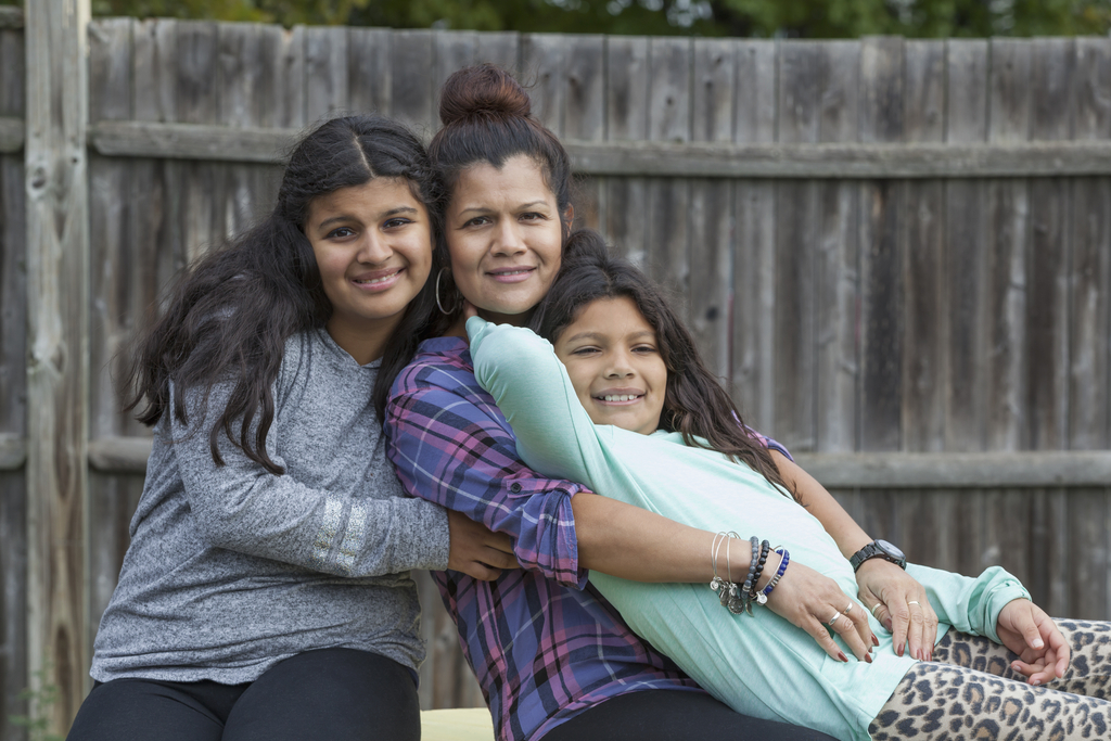 A mother and two daughters embrace  outdoors near a fence.