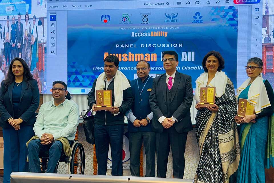 Dr. Mitra and fellow panelists smiling. The panelists hold congratulatory plaques and stand in front of a slide show that reads: AccessAbility Panel Discussion: Ayushman for All.