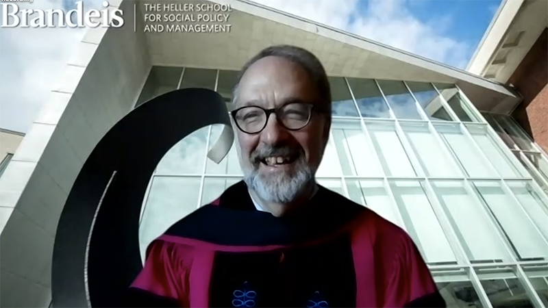 Dean David Weil in red graduation regalia in front of a Zoom background of the sweeping windows of the Zinner Forum 