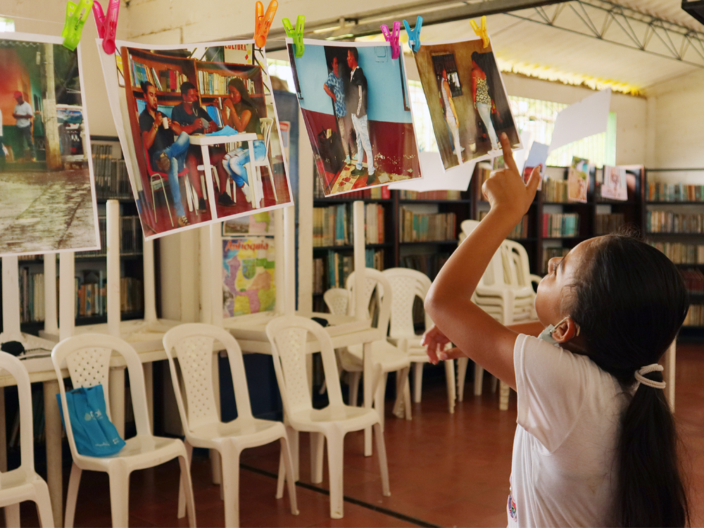 Slide 3 of 7 - A girl points at a series of printed photos hanging on a string in a classroom, each photo depicting multiple community members in different spaces, in Urama, Colombia.