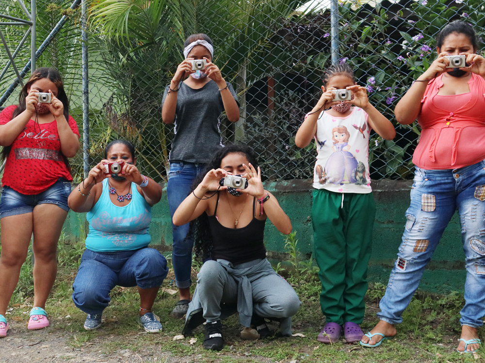 Slide 2 of 7 - Six young women, some standing and some crouching, point cameras toward the viewer, in Cruces, Colombia.