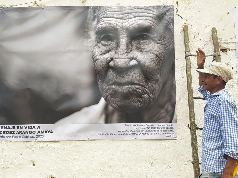 Slide 5 of 7 - A man with a ladder smiles while looking at a poster with a photograph of an older man, in Urama, Colombia.