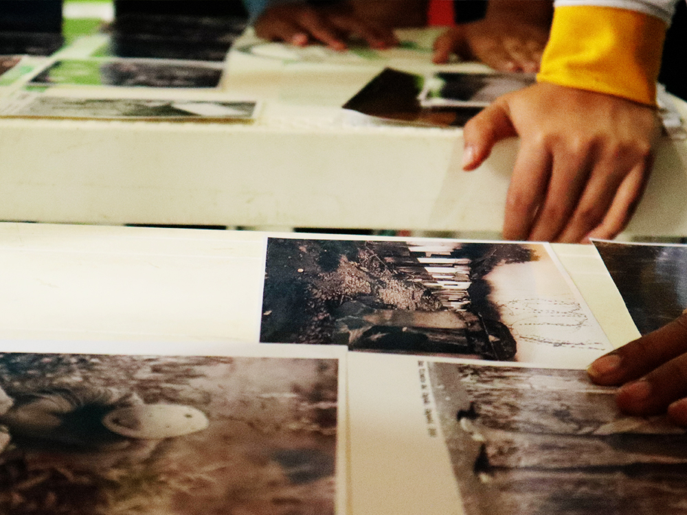 Slide 4 of 7 - A close-up of hands on a table as people look over black and white photographs, in Cruces, Colombia.