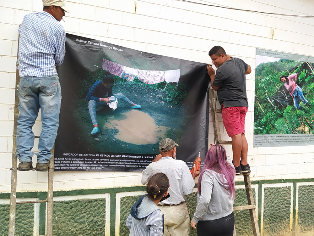 Slide 6 of 7 - Two men stand on ladders to hang a poster, while three other people help. The poster depicts a woman sitting next to a muddy hole in the road, with laundry hanging behind her, in Urama, Colombia.