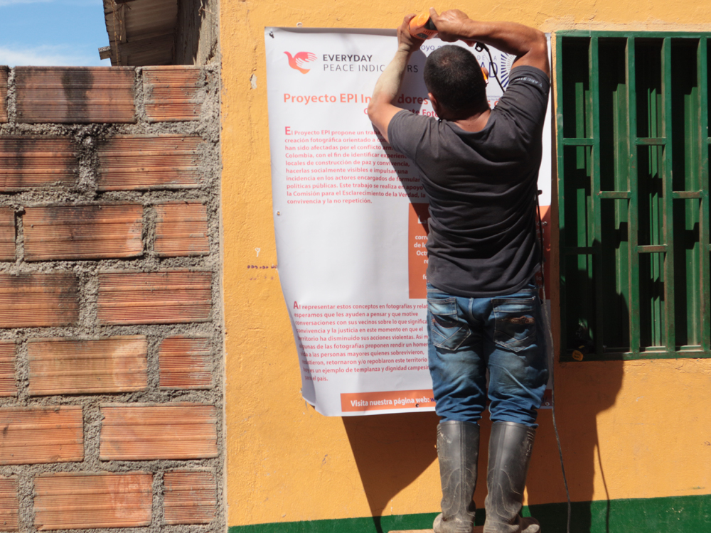 Slide 7 of 7 - A man uses a drill to install a poster explaining the photovoice project and Everyday Peace Indicators, in Cruces, Colombia.